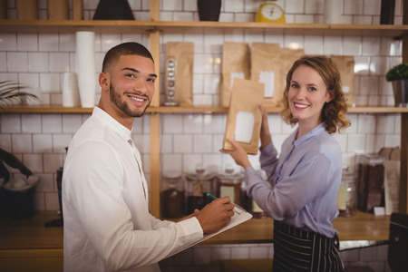 Portrait of smiling male owner and waitress arranging packages on shelf at coffee shopの写真素材