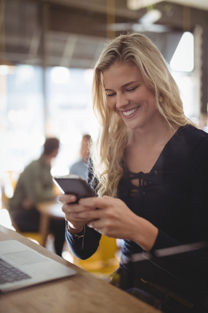 Smiling young blond woman texting through mobile phone while sitting at counter in coffee shopの写真素材