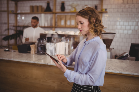 Young pretty waitress using digital tablet while standing by counter at coffee shopの写真素材