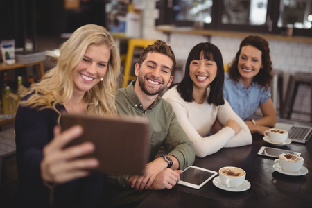 Young woman taking selfie with friends from tablet while sitting at table in cafeの写真素材