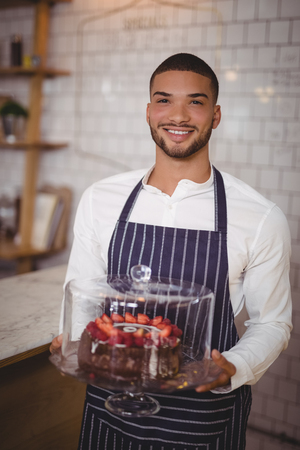 Portrait of smiling young waiter holding cake on glass cakestand at coffee shopの写真素材