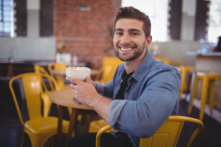 Portrait of smiling handsome man holding fresh coffee cup while sitting on chair at cafeの写真素材