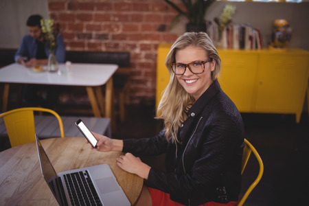 Portrait of smiling young blond woman holding smartphone while sitting with laptop at table in coffee shopの写真素材