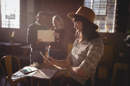 Smiling woman using smartphone against colleauges standing at coffee shopの写真素材