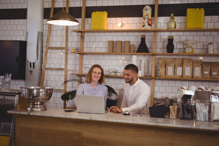 Young male owner and waitress using laptop while sitting at counter in coffee shopの写真素材
