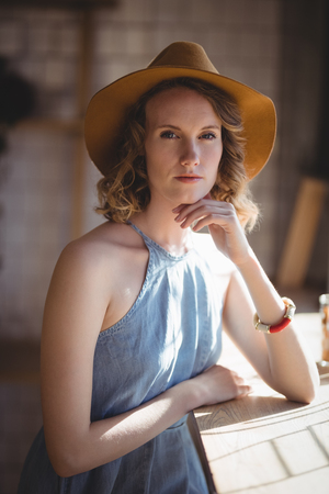 Portrait of beautiful young woman wearing hat standing by table at coffee shopの写真素材