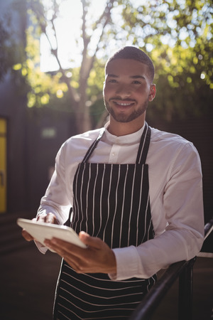 Portrait of smiling young waiter using digital tablet while leaning on railing during sunny dayの写真素材