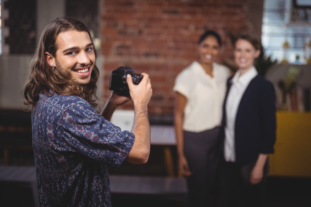 Portrait of smiling young photographer photographing female colleagues at coffee shop