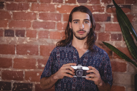 Portrait of confident young photographer holding camera against brick wall at coffee shopの写真素材