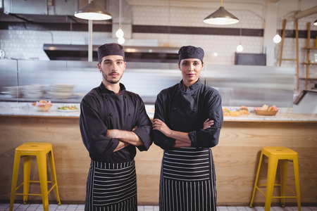 Portrait of confident young wait staff in black uniform standing with arms crossed against counter in coffee shopの写真素材