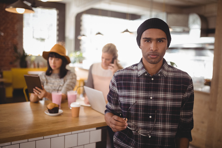 Portrait of young man holding eyeglasses standing against female friends sitting at table in coffee shopの写真素材