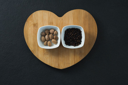 Nutmegs and black pepper seeds in heart shaped chopping board on black backgroundの写真素材
