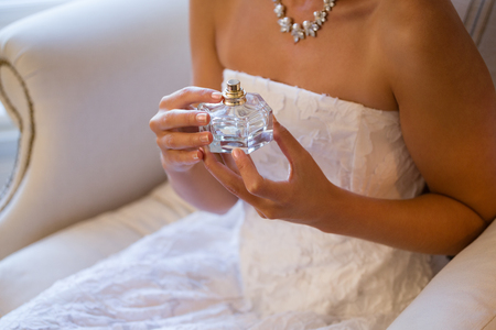 Midsection of bride holding perfume sprayer while sitting on armchair at homeの写真素材