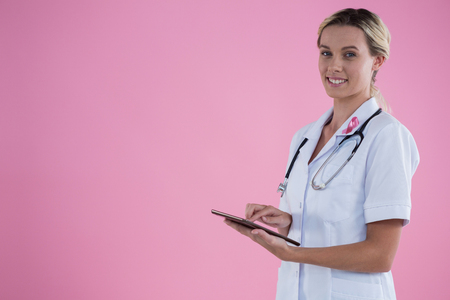 Portrait of smiling female doctor holding tablet computer while standing against pink backgroundの写真素材