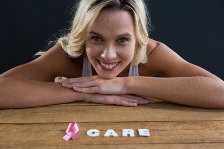 Portrait of smiling woman leaning on table with pink ribbon and care text against black backgroundの写真素材