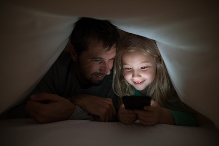 Father and daughter lying under blanket and using mobilephone at homeの写真素材