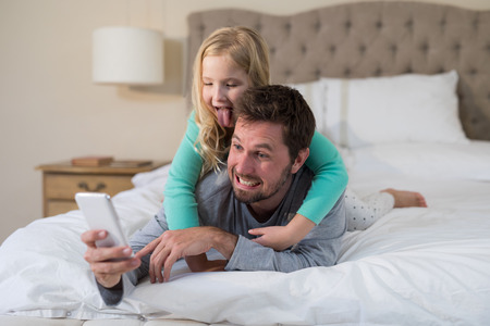 Father and daughter taking a selfie on bedの写真素材