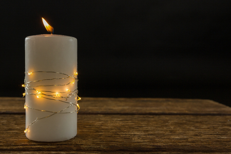 Illuminated string lights wrapped on lit candle at table against black backgroundの写真素材
