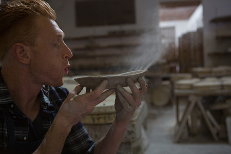 Male potter blowing dust from a bowl in pottery workshopの写真素材