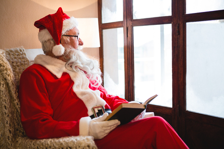 Thoughtful santa claus reading novel in living room during christmas timeの写真素材