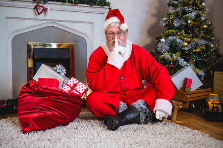 Santa claus with gift sack sitting on rug with finger on lips in living room at home