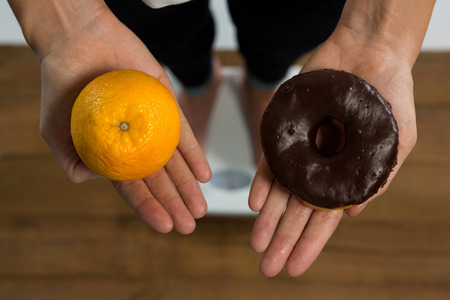 Close-up of woman standing on weight machine and holding doughnut and orange
