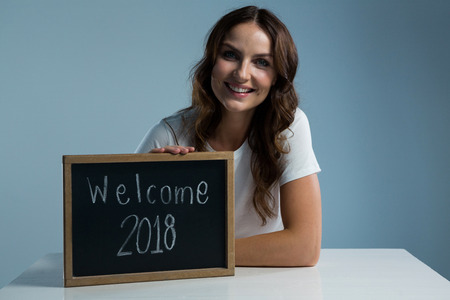 Portrait of smiling woman showing slate with text welcome 2018 against grey backgroundの写真素材