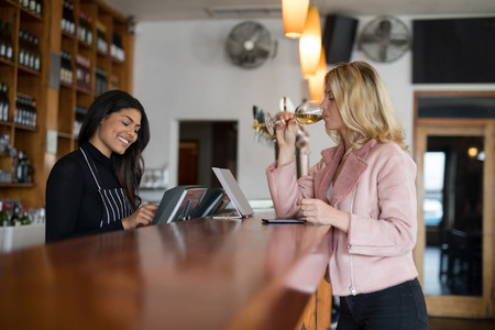 Waitress standing at counter while woman having glass of wine in barの写真素材