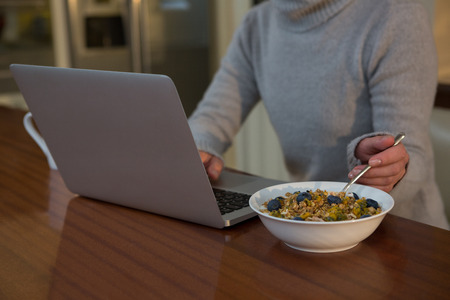 Mid section of woman having breakfast while using laptop at homeの写真素材