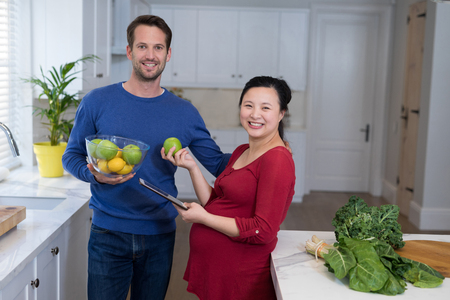 Smiling pregnant couple holding sweet limes and digital tablet in the kitchenの写真素材