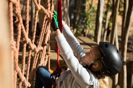 Close-up of little girl wearing helmet climbing on rope fence in the forestの写真素材