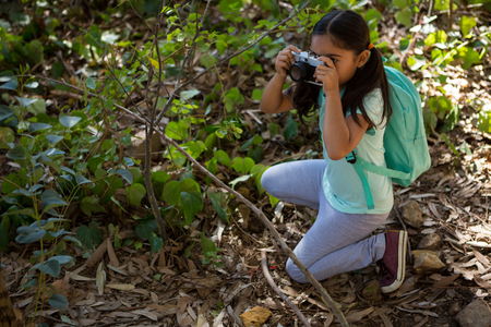 Little girl with backpack taking photo with camera on a sunny day in the forestの写真素材