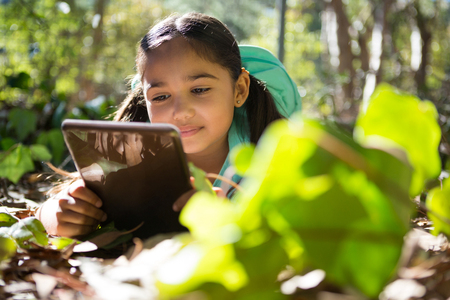 Little girl with backpack lying on ground using her tablet on a sunny day in the forestの写真素材