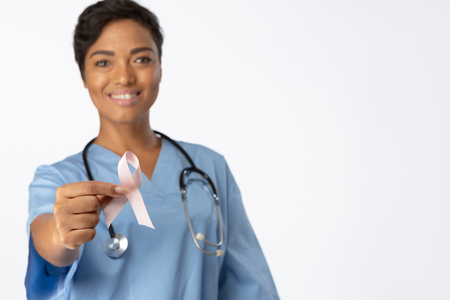 Smiling nurse holding breast cancer awareness pink ribbon between her fingers on a white foregroundの写真素材