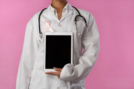 Nurse wearing pink ribbon holding a tablet in her hand on a pink background for breast cancer awarenessの写真素材
