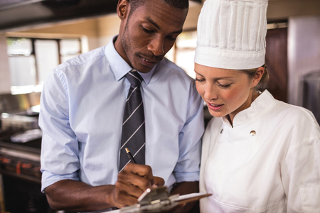 Male manager and female chef writing on clipboard in kitchen at hotelの写真素材