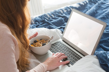 Woman using laptop while having breakfast in bedroom at homeの写真素材