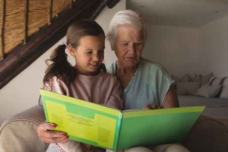 Front view of a grandmother and granddaughter reading story book in living room at homeの写真素材