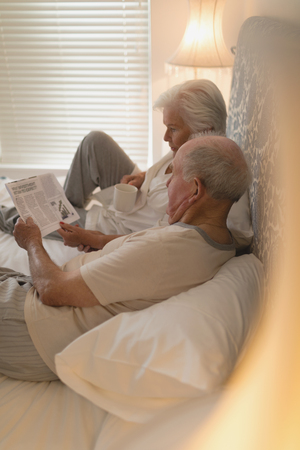 Side view of senior couple reading newspaper and drinking on the bed at homeの写真素材