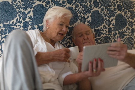Front view of senior couple using digital tablet and drinking in the bed at homeの写真素材