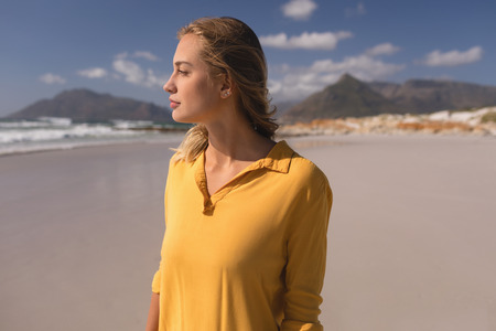 Thoughtful young woman standing on the beach against mountains in backgroundの写真素材