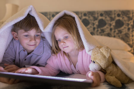 Front view of cute siblings using digital tablet under blanket in bedroom at homeの写真素材