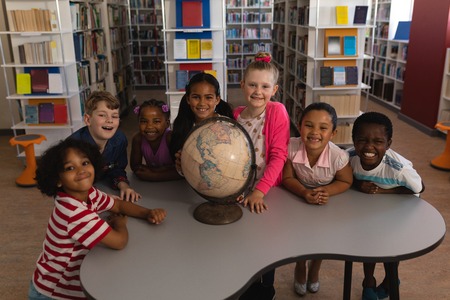 Front view of happy schoolkids with globe looking at camera in school libraryの写真素材