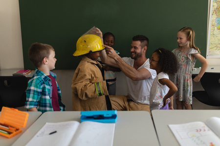 Front view of male Caucasian firefighter helps to wearing fire uniform to schoolkid with all the schoolkids around in classroom of elementary schoolの写真素材