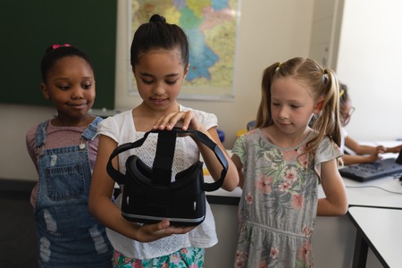 Front view of schoolgirl with curious classmates holding a virtual reality headset in classroom of...