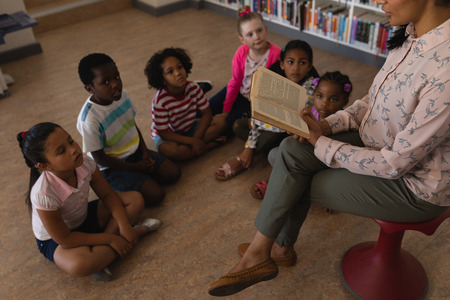 High angle view of female teacher sitting on chair and reading a story schoolkids sitting on floor in school libraryの写真素材