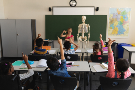 Rear view of schoolkids raising hands and sitting at desk in classroom of elementary schoolの写真素材