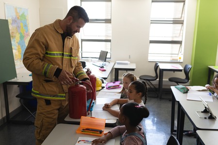 Side view of male Caucasian firefighter teaching schoolkids about fire safety, showing extinguisher in classroom of elementary schoolの写真素材
