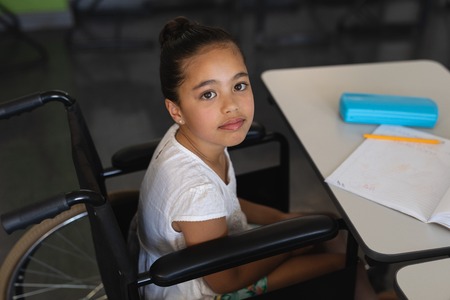 High angle view of disabled schoolgirl looking at camera and sitting at desk in classroom of elementary schoolの写真素材