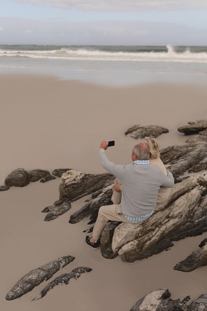 Front view of active Senior couple taking selfie with mobile phone standing on rocks at the beach...